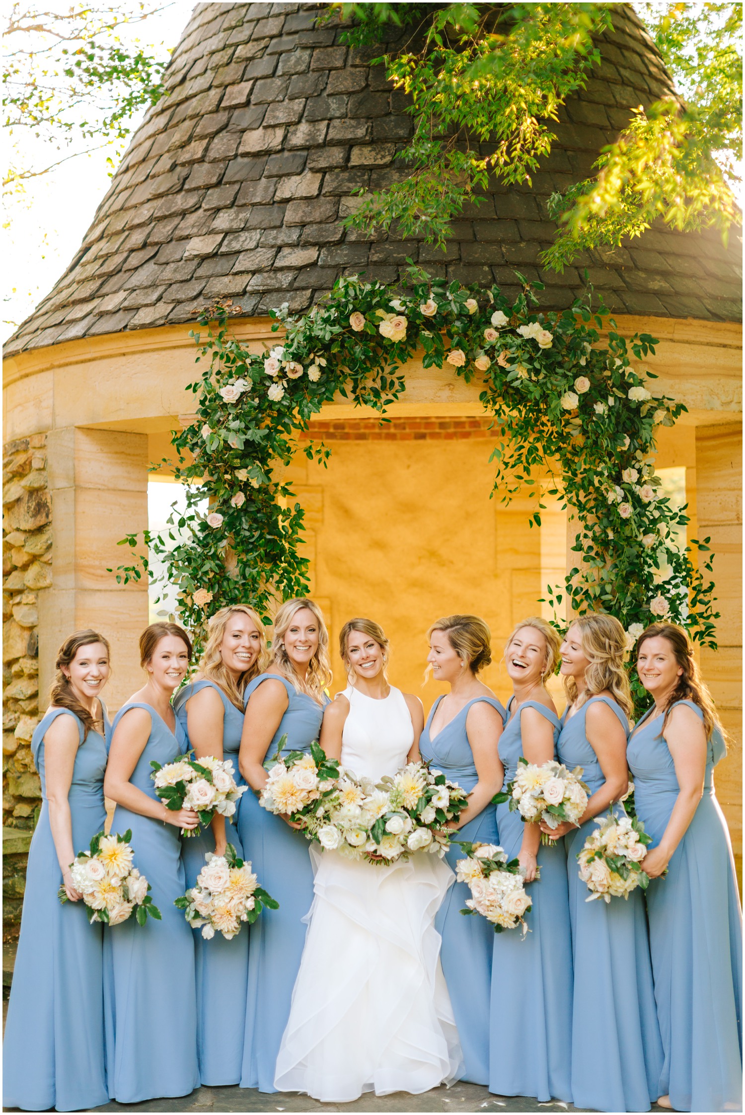 bride poses with bridesmaids under floral arch at Graylyn Estate