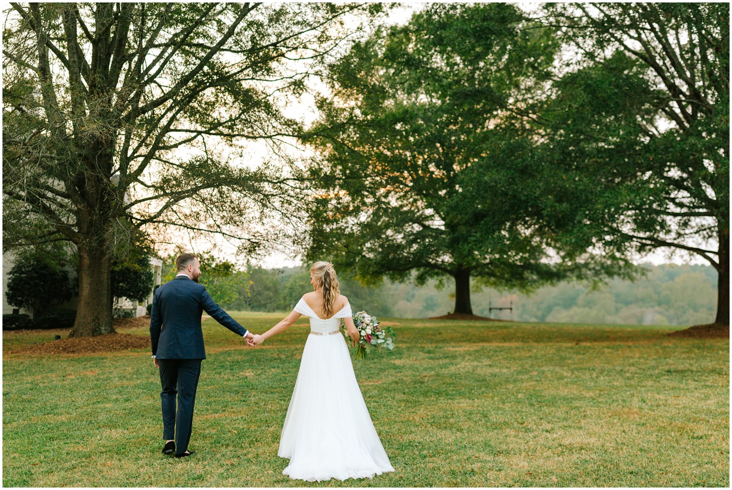 bride and groom hold hands walking through field 