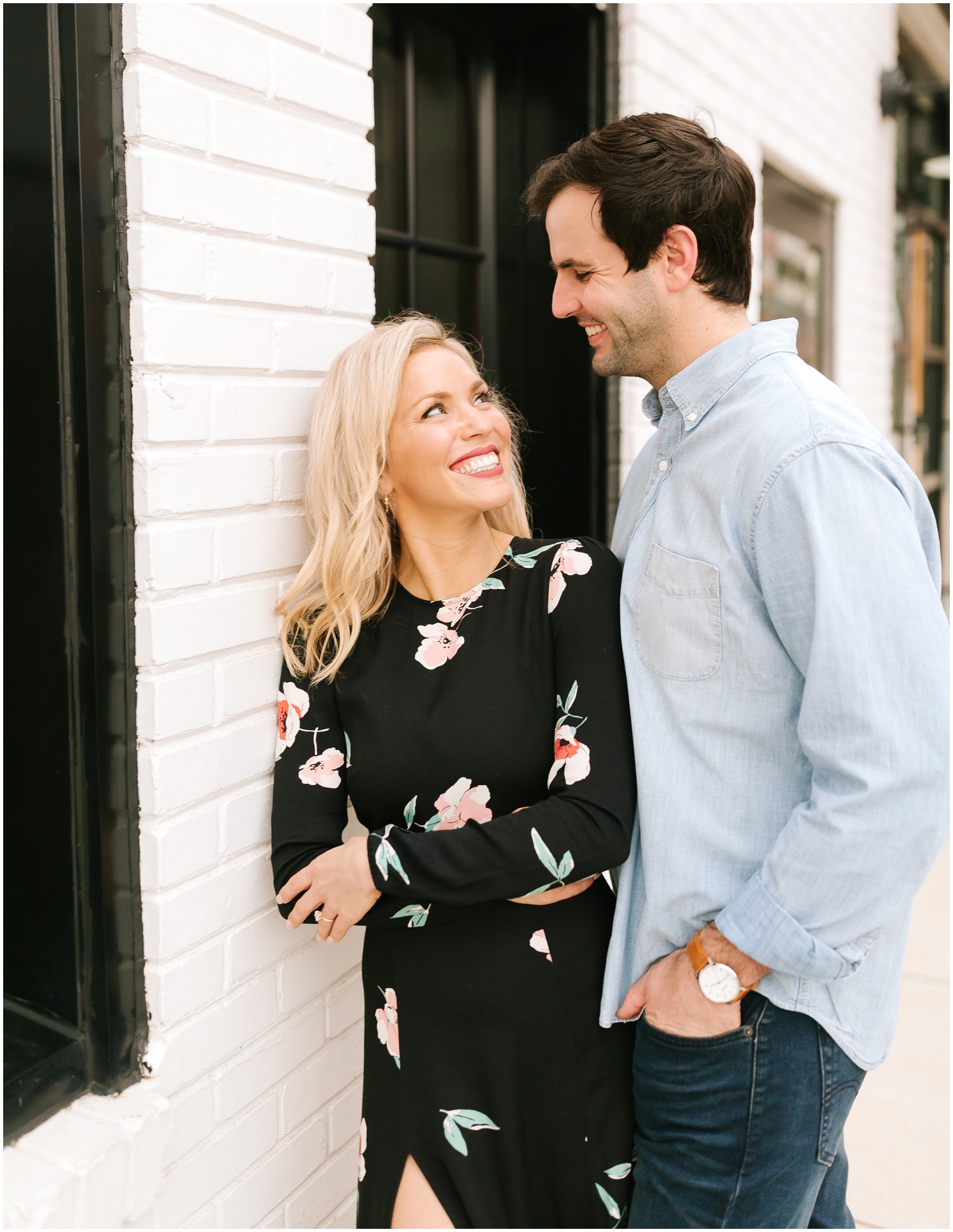bride wears black gown with flowers and looks at groom in blue button up shirt during engagement portraits