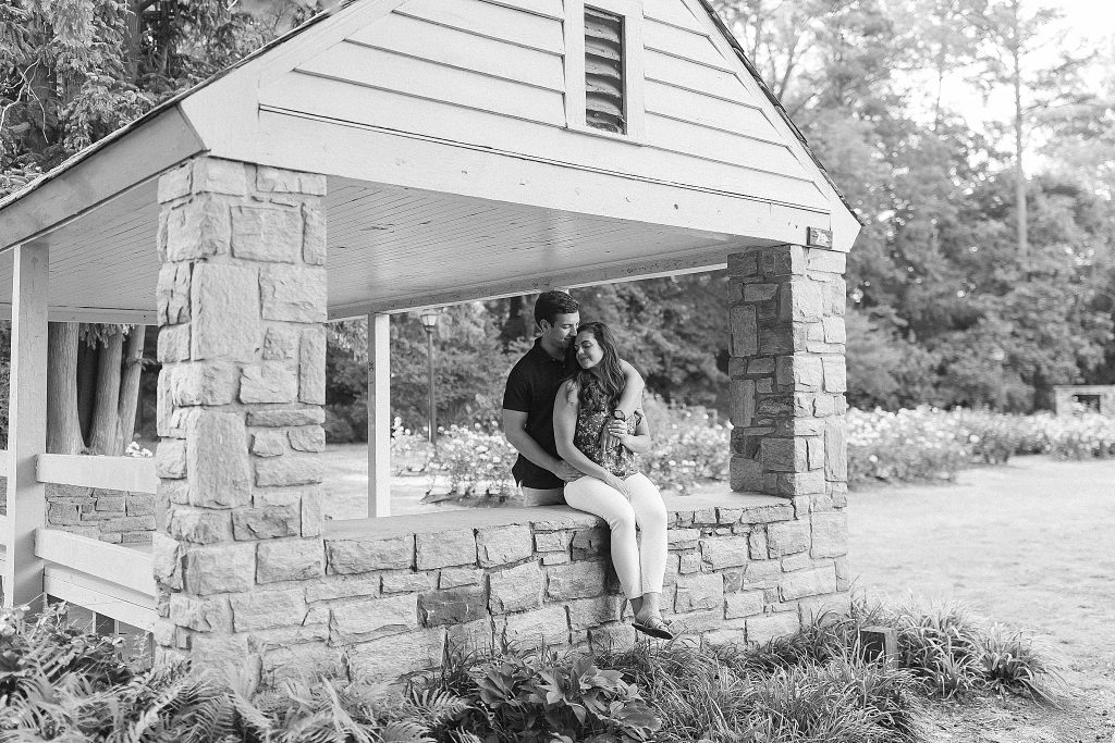 black and white portrait of engaged couple sitting on brick wall at Raleigh Rose Garden