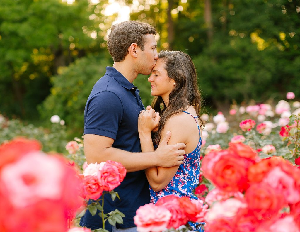 groom to be kisses bride to be on forehead while standing in pink rose bush