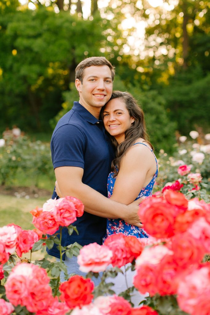 engaged couple poses next to pink roses at Raleigh Rose Garden