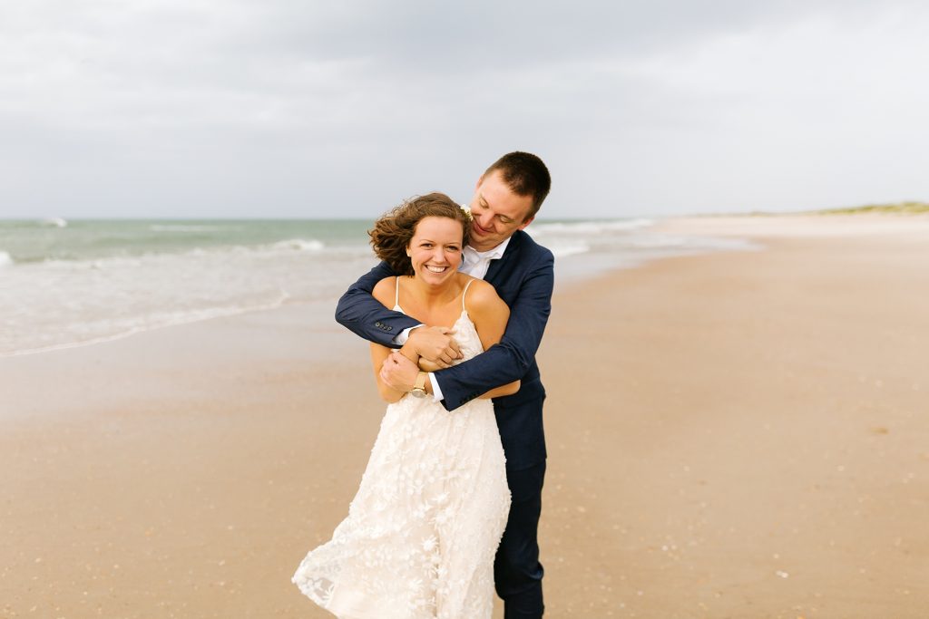 groom hugs bride from behind during wedding photos on beach