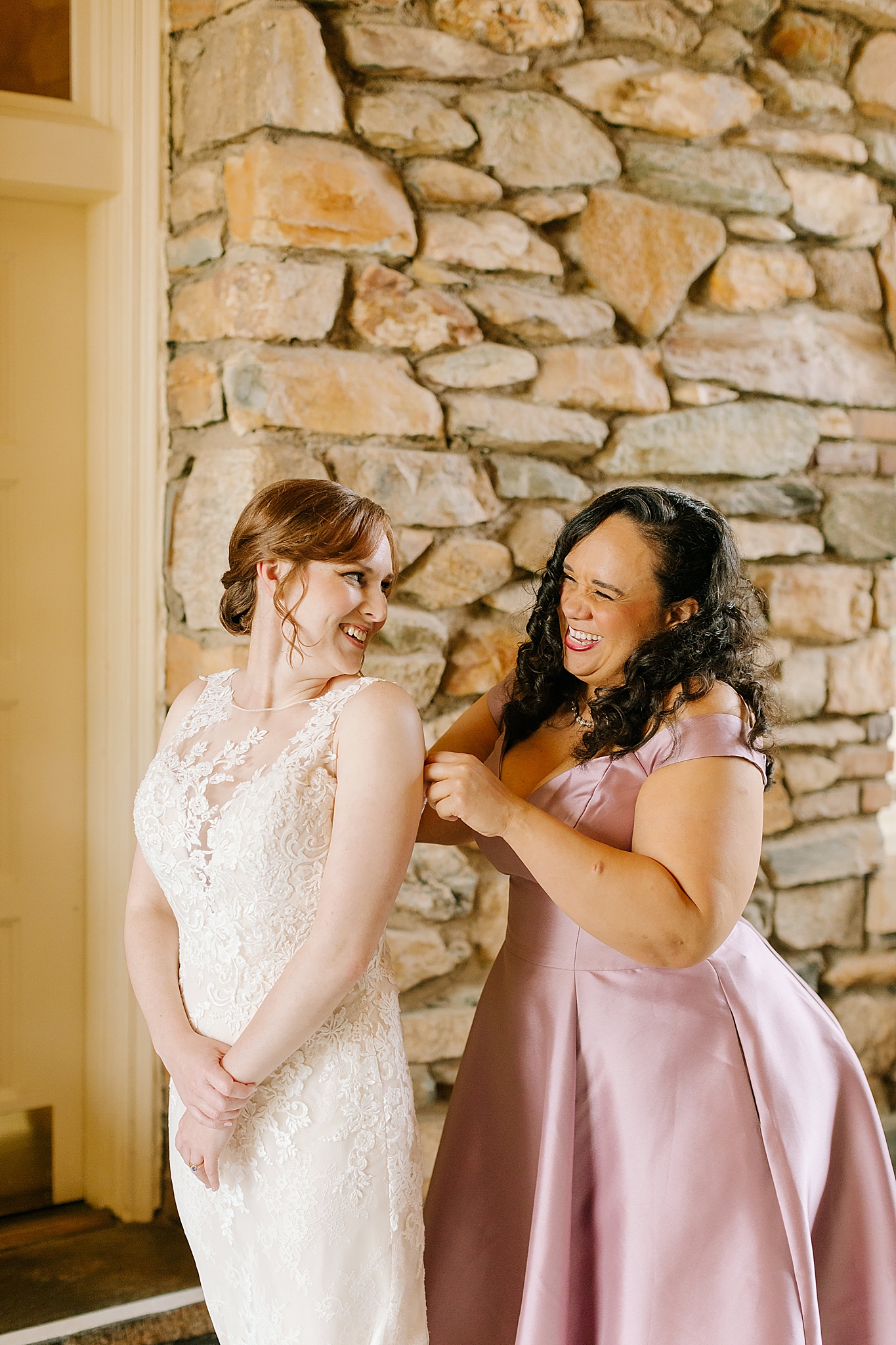 bridesmaid in pink satin gown helps bride with wedding dress