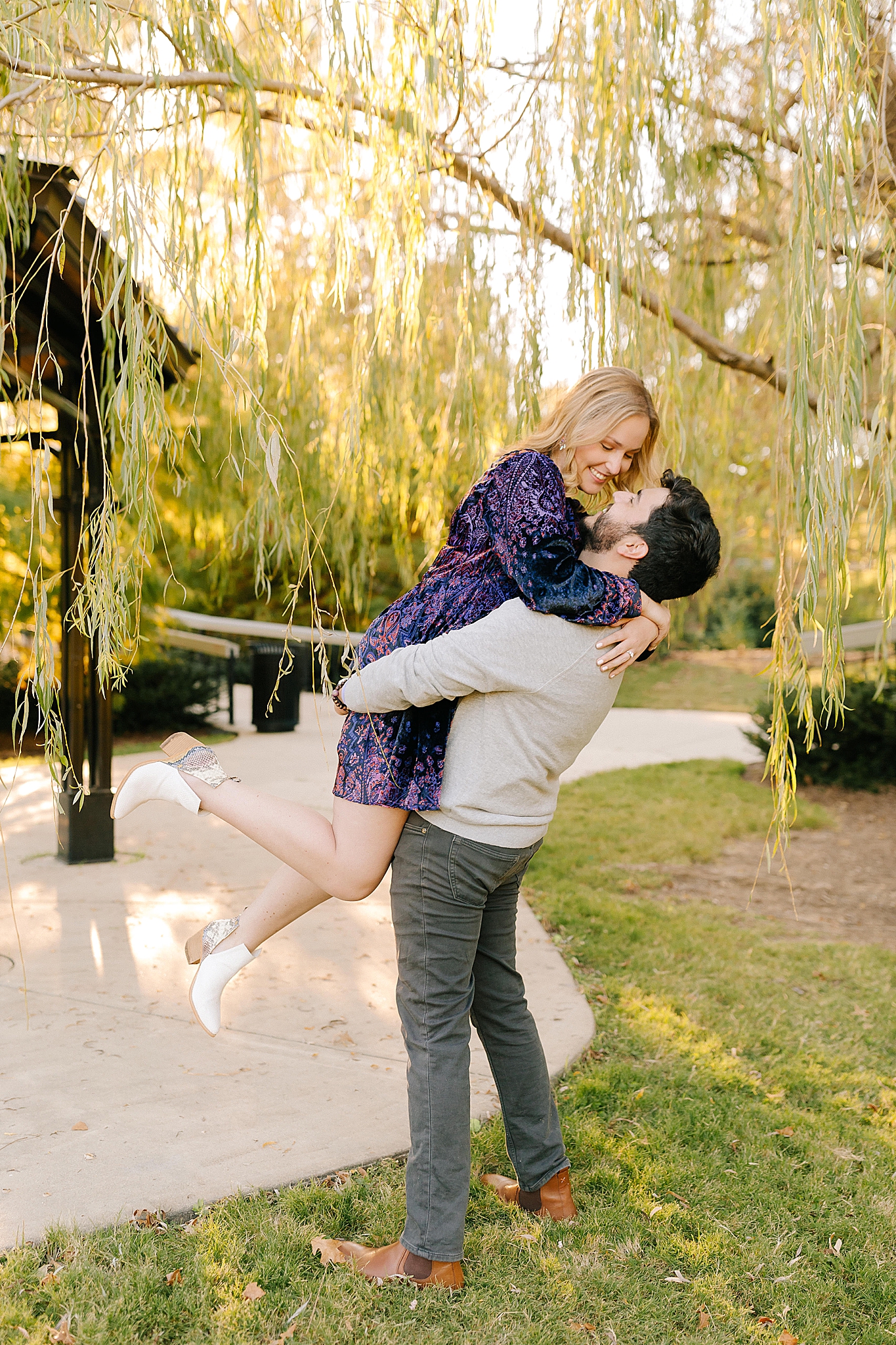 groom lifts bride during engagement photos by pavilion in Pullen Park