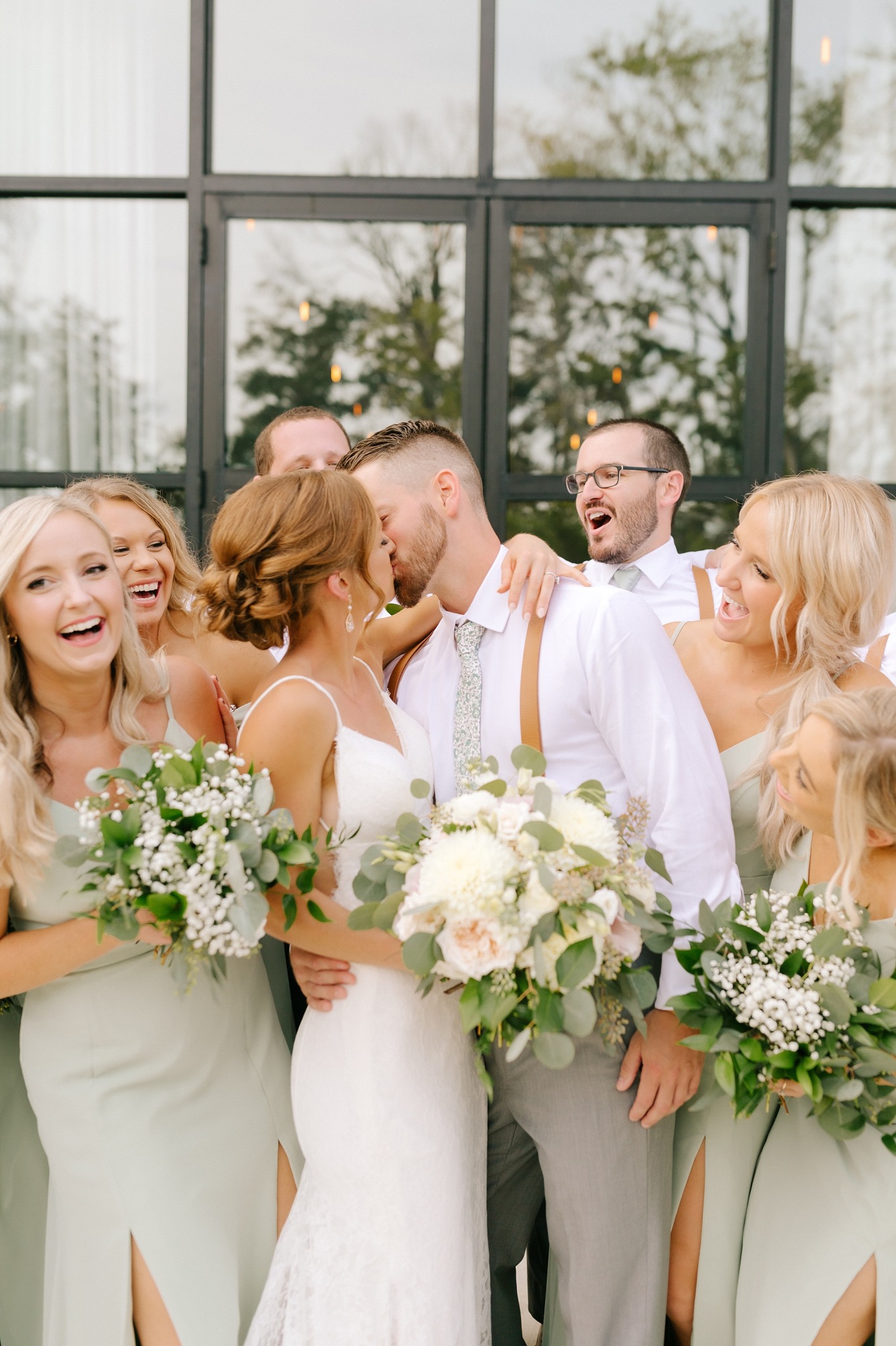 bride and groom kiss with bridesmaids laughing around them