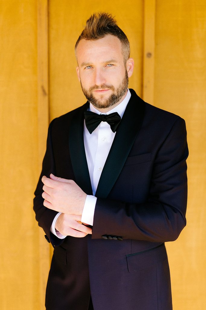 portrait of groom in tux in front of bright yellow wall