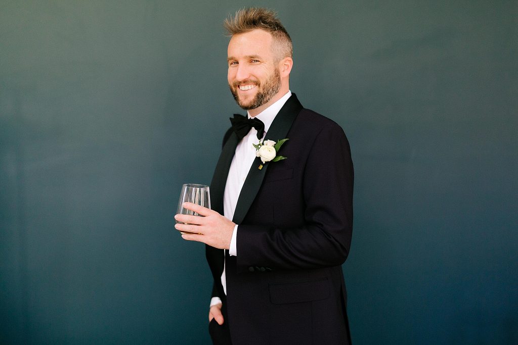 groom holds glass and poses by teal wall