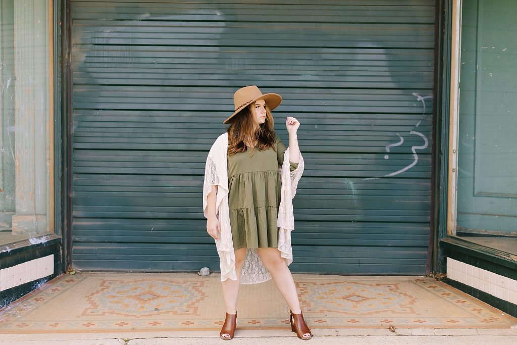 senior with wide brimmed hat and olive green dress poses in Downtown Winston-Salem