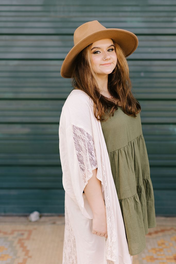 senior girl in green dress with white sweater poses by teal garage 