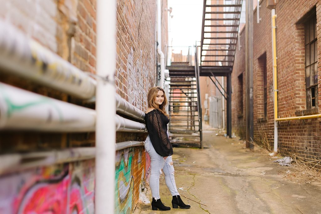 senior in black top and jeans leans against pipes on wall in Downtown Winston-Salem