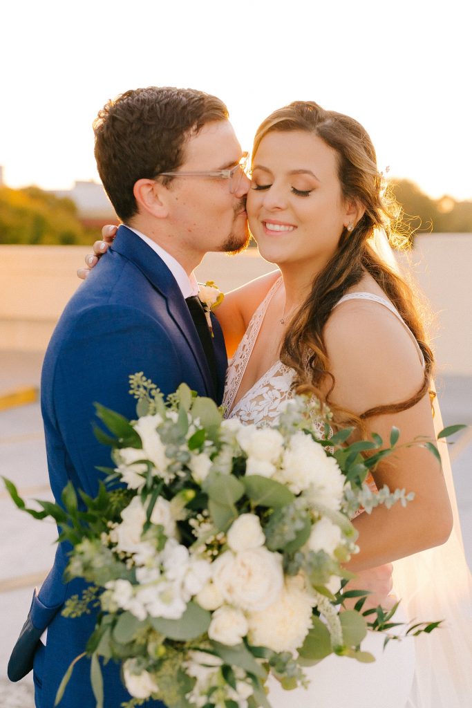 groom kisses bride's cheek during sunset wedding portrait on roof