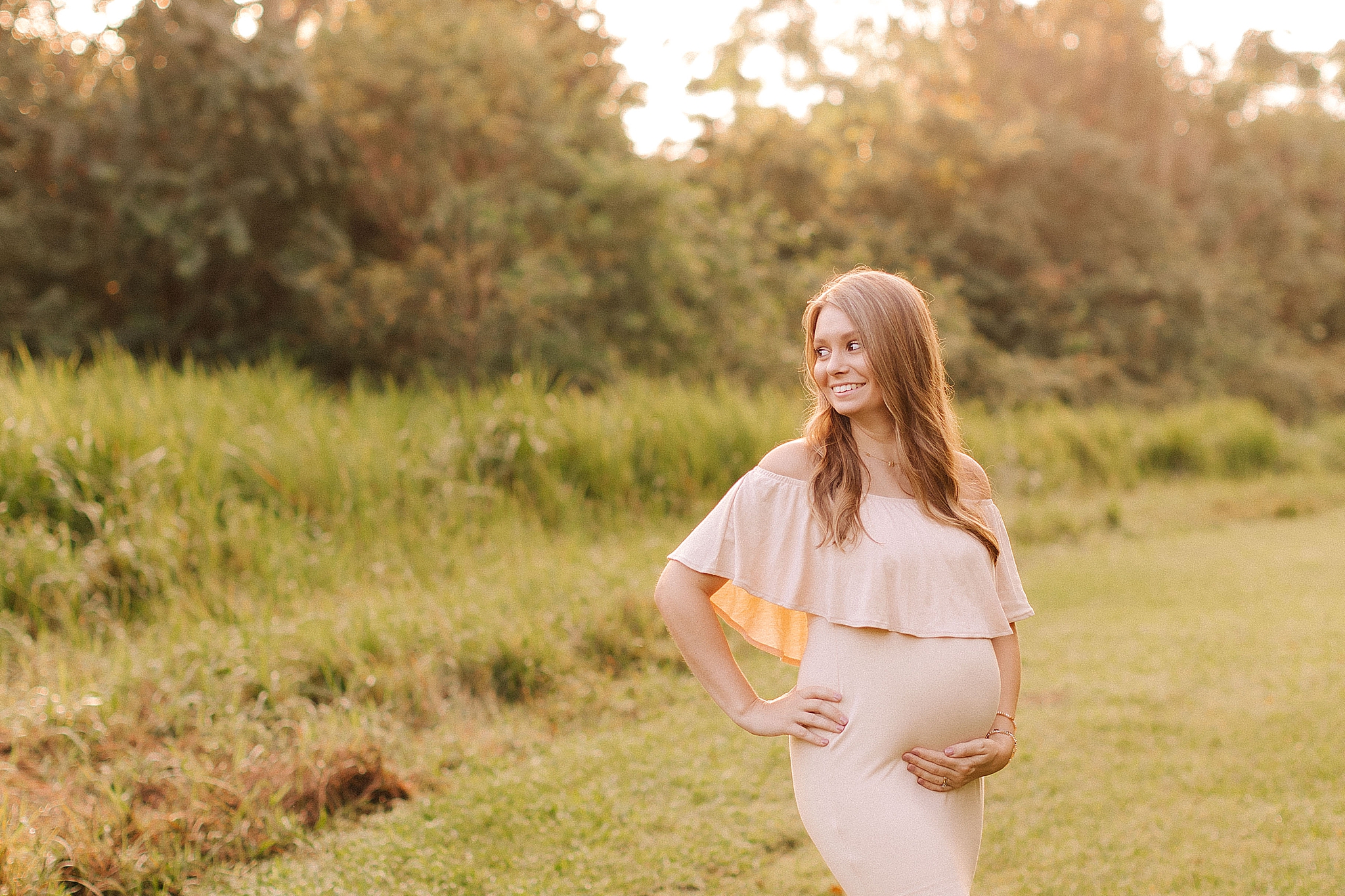 mom in pale pink gown holds baby belly standing on edge of field