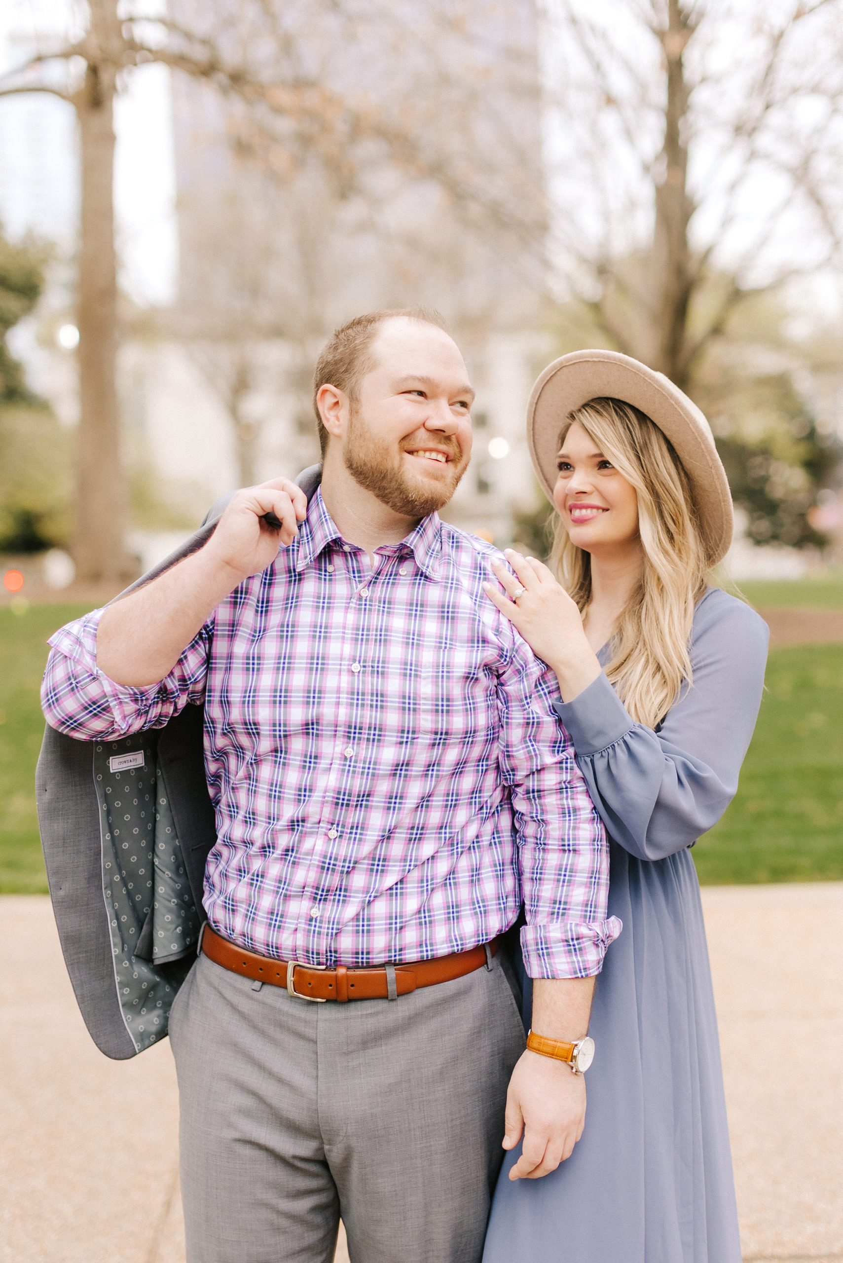 bride in wide brimmed hat looks at groom during Downtown Raleigh engagement photos 