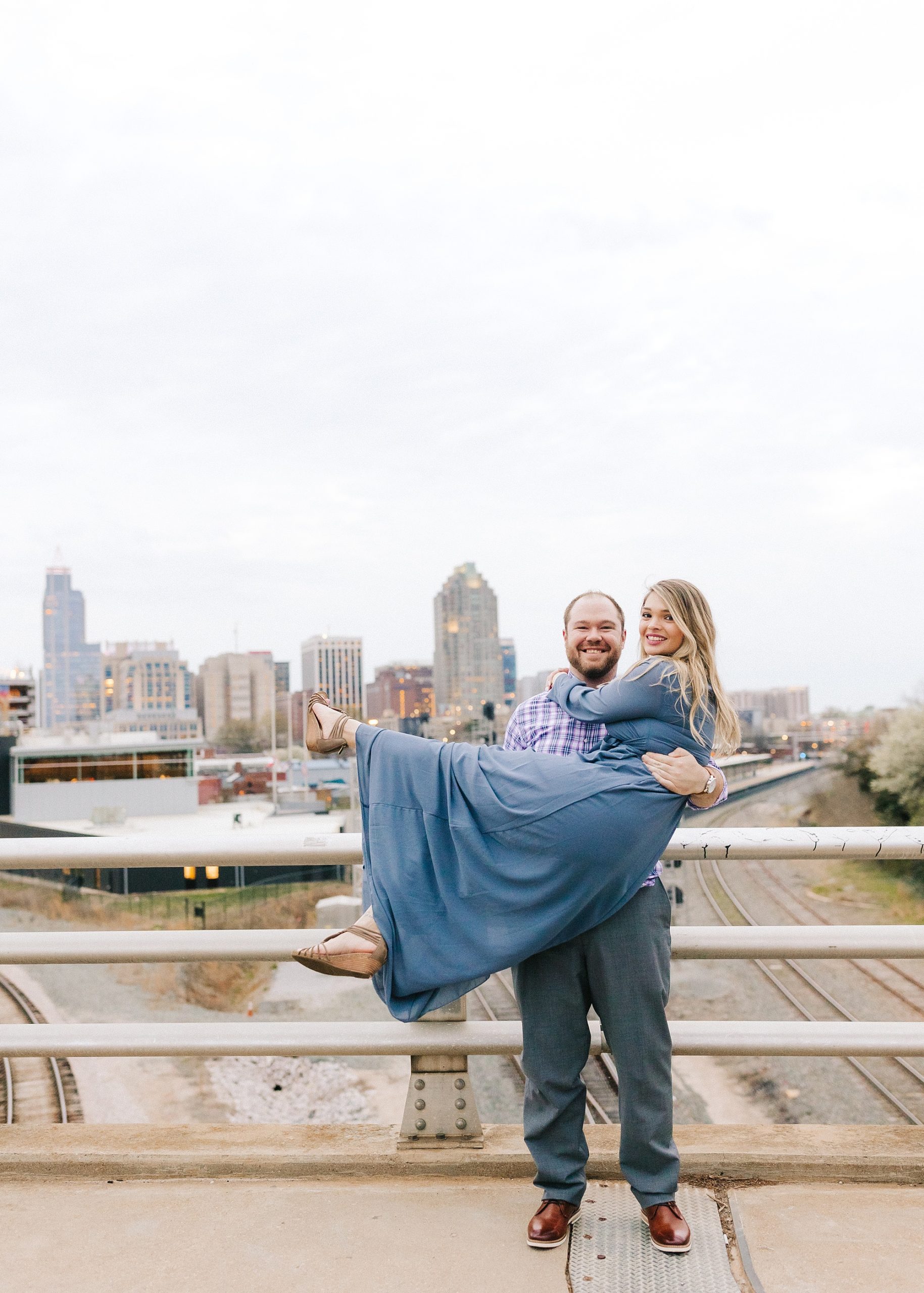 groom lifts bride up in front of Raleigh skyline