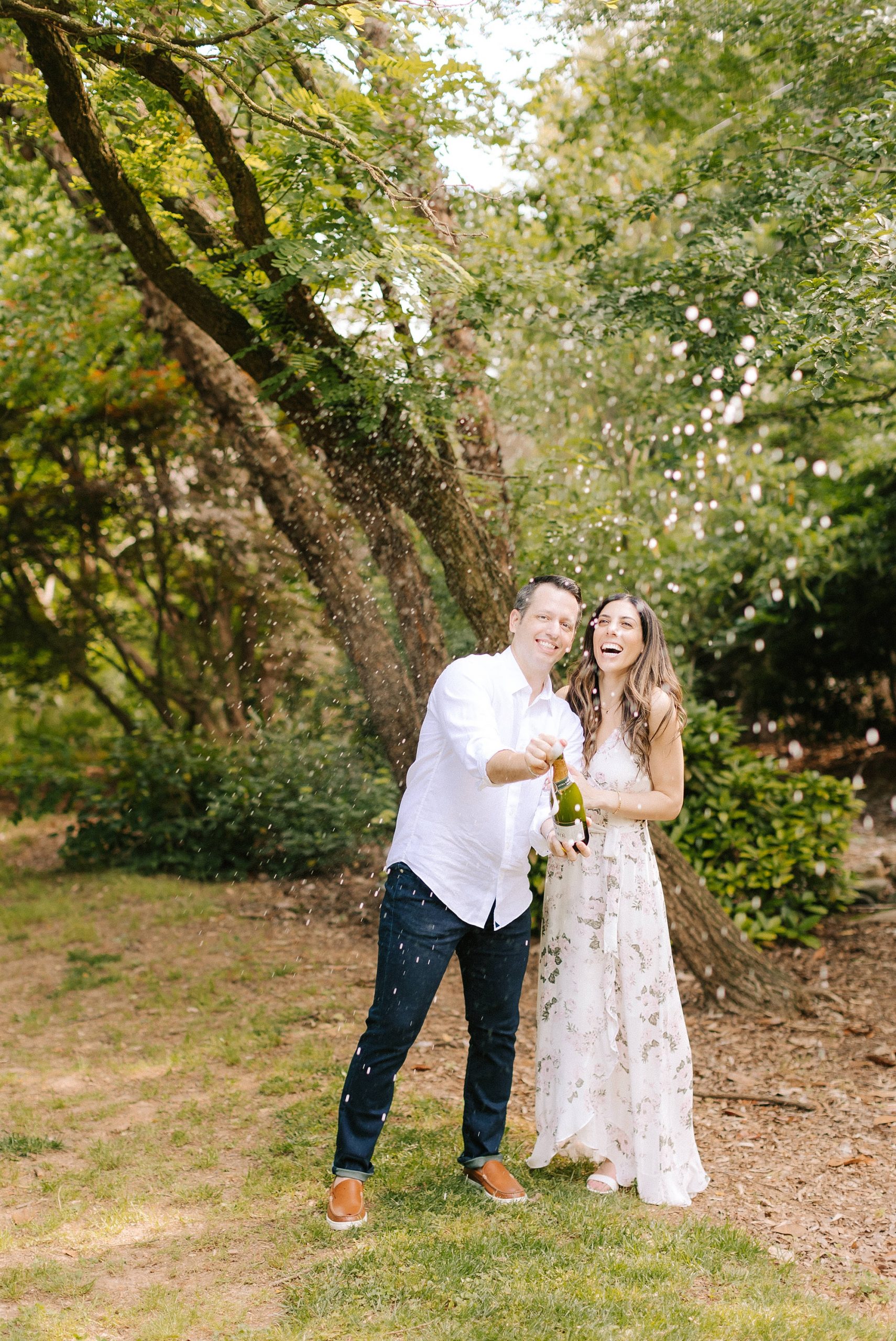 bride and groom pop champagne during JC Raulston Arboretum engagement session