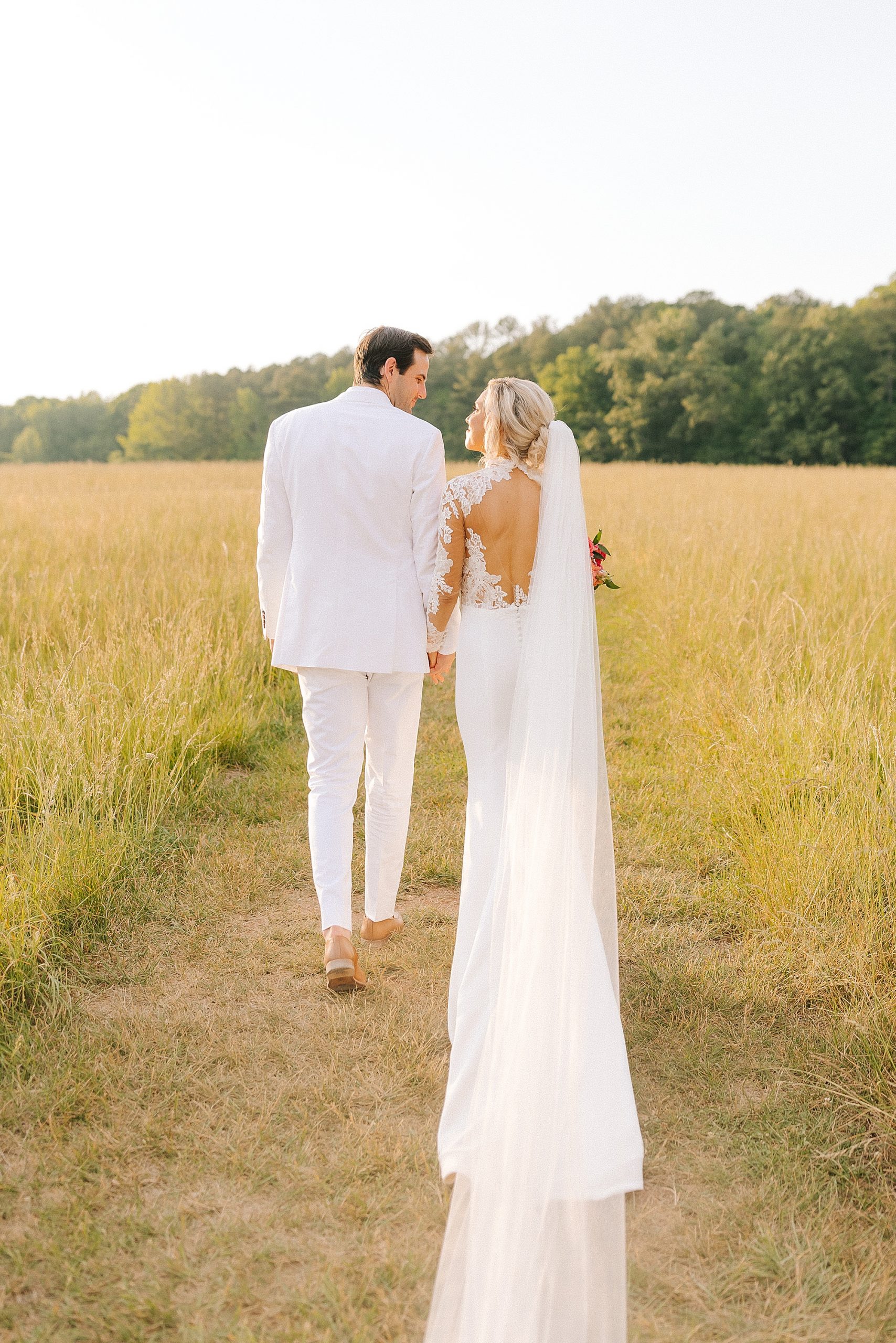 newlyweds walk through path in field at The Meadows Raleigh with bride's veil trailing behind her 