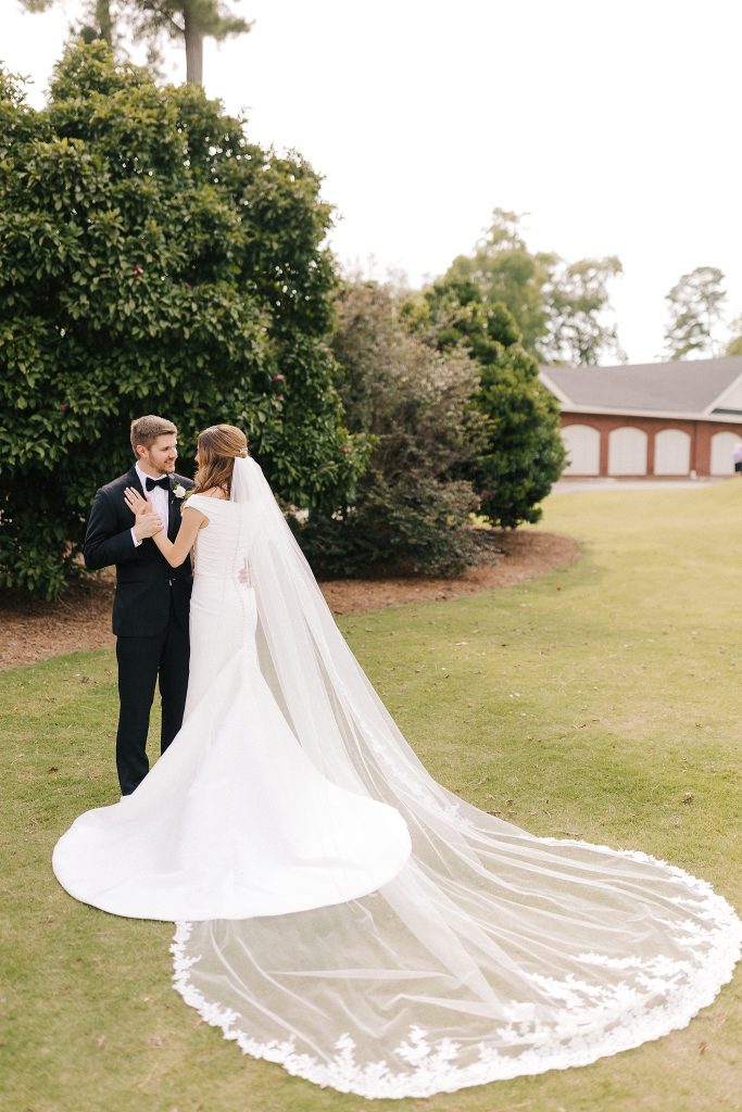bride and groom pose outside Prestonwood Country Club with bride's veil behind her 
